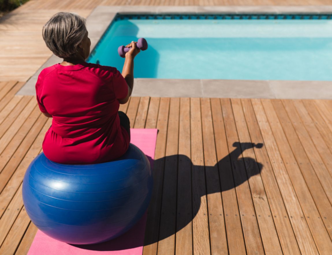 Rear view of senior African American woman exercising with dumbbells on a swiss ball in front of the swimming pool in the backyard of home