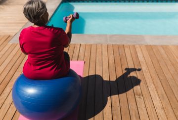 Rear view of senior African American woman exercising with dumbbells on a swiss ball in front of the swimming pool in the backyard of home