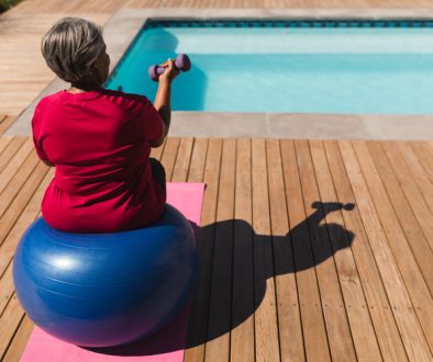 Rear view of senior African American woman exercising with dumbbells on a swiss ball in front of the swimming pool in the backyard of home