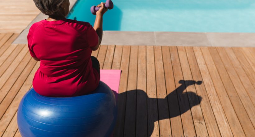 Rear view of senior African American woman exercising with dumbbells on a swiss ball in front of the swimming pool in the backyard of home